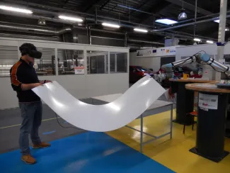 A human and a collaborative robot are transporting a large flexible sheet of plastic towards a table. 